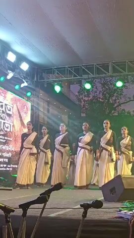 A group of seven women on a stage are performing a dance routine. They are dressed in traditional Indian clothing, specifically sarees. The stage is lit with bright lights, and there is a large screen in the background displaying text and images in Bengali. Microphones are set up in the foreground, indicating that the performance is likely part of a larger event such as a festival or cultural celebration. The women are synchronized in their movements, suggesting a choreographed performance. The overall atmosphere appears to be that of a live event.