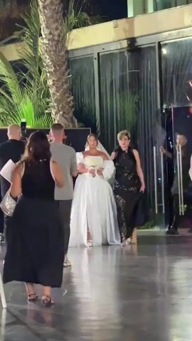 The video shows a wedding procession with a bride in a white gown walking forward, accompanied by two women and two men. The bride is holding a bouquet of white flowers and is wearing a veil. One woman to her right wears a black sparkly dress, and another woman in a black dress is walking in front of her, facing away from the camera. Two men, one in a black shirt and pants, holding a camera, and another in a grey shirt and black pants, are walking beside or in front of the bride, appearing to be part of the wedding party or event staff. The scene appears to be outdoors at night, possibly at a wedding reception or ceremony venue, with palm trees and modern building architecture visible in the background. The lighting suggests it is evening or nighttime. The overall atmosphere seems celebratory, given the context of a wedding. There is no visible text in the video.