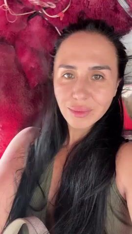 The video begins with a close-up of a woman's face. She has long, dark hair and is wearing a green tank top. She is smiling and looking directly at the camera. The background is blurred, but it appears to be the interior of a vehicle. As the video progresses, the camera moves slightly, revealing more of the interior. The vehicle appears to be a tuk-tuk, with red, fuzzy upholstery lining the roof and sides. Through the windows, a blurry outdoor scene can be seen, suggesting the vehicle is in motion. The woman continues to look at the camera and smile throughout the short clip.