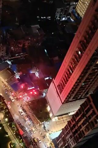 This video is an aerial view of a city street at night. A tall, modern building dominates the right side of the frame, with its lights illuminated. Below and to the left of the tall building, several lower buildings are visible, some with blue roofs. The street in front of these buildings is lined with parked cars and motorcycles, and a stream of traffic moves along it. Red and white lights are visible, possibly from emergency vehicles or temporary construction lighting. Several bursts of bright light, appearing to be fireworks, illuminate the scene from below. People are gathered on the street, observing the fireworks. The overall atmosphere is one of a lively urban night scene with a celebratory event.