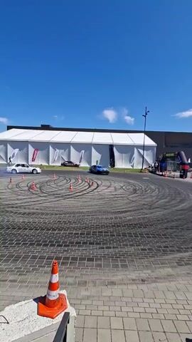 Two drift cars are performing donuts on a paved lot in front of a large white tent structure. The sky is blue with a few white clouds. Orange traffic cones are scattered around the lot. One car, blue and purple with the words "TRANS AGENT" on its side, is in the foreground. The second car, white with racing stripes and the words "ATOMBO" on its side, is in the background, drifting in a wider circle. Both cars are kicking up smoke from their tires. Several spectators can be seen in the background near a tent with an inflatable archway.