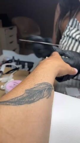 A close-up shot focuses on a person's forearm with a feather tattoo as their hand is being worked on by a manicurist. The manicurist, wearing black gloves and a striped top, is using a nail file on the client's fingernails. Various nail care products and tools are visible on a surface in the background, suggesting a nail salon environment.