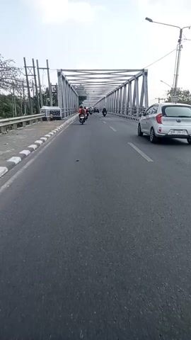 A short clip shows a camera mounted on a vehicle moving across a bridge. The bridge has a metal truss structure and a paved road. Several motorcycles and a car are visible on the bridge, all moving in the same direction as the camera. The weather appears to be cloudy. The camera angle is slightly tilted.