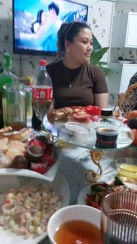 The video shows a gathering of people around a table laden with food, likely a meal or celebration. In the foreground, there are various dishes, including what appears to be a salad, cupcakes, and a platter with meat, sausage, and sliced tomatoes. A bottle of Coca-Cola is visible on the left side of the table. In the middle ground, a woman in a brown t-shirt is seated, and a woman in a patterned shirt is laughing and holding her hand to her mouth. A fruit basket filled with bananas, plums, and apples sits on the table. Further back, another woman in a pink top is seated and holding a glass. The environment appears to be indoors, with a television screen displaying an image of a couple in the upper left corner of the frame. The lighting is warm, suggesting an evening setting. The overall atmosphere seems festive and convivial.