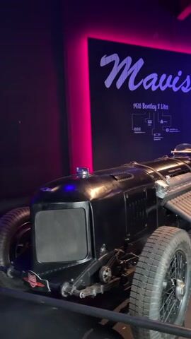 A black vintage race car is on display in a dimly lit room. The car is a 1930 Bentley 8 Liter and is named "Mavis." The car has a large engine with visible pipes and wires. The wheels are spoked and the tires are thick. The car is positioned on a slight incline. A black wall behind the car is illuminated with pink and blue lights. Text on the wall reads "Mavis" in large script, and smaller text below states "1930 Bentley 8 Liter" along with diagrams. To the right, more text lists names and technical specifications for other vehicles. The car is roped off with a thin metal barrier.