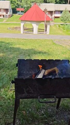 This media file shows a close-up of a metal barbecue grill with burning logs and smoke. In the background, a red-roofed gazebo with arched openings stands in a grassy area. Beyond the gazebo, several small buildings with light-colored exteriors and elevated entrances are visible, suggesting a resort or a dacha complex. The setting appears to be outdoors on a sunny day, with green grass and trees. The overall atmosphere is peaceful and suggests an outdoor leisure activity like a picnic or barbecue. There is no visible text in the media file.