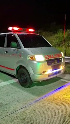 A silver ambulance is parked on a paved surface at night, facing slightly to the right of the frame. Red flashing lights are mounted on its roof. The word "AMBULANCE" is visible in reverse on the front grille and correctly on the side of the vehicle. A red heartbeat graphic is also on the side. The ambulance's headlights are on, and multicolored emergency lights (blue and yellow/orange) are reflecting on the ground in front of it. Some dark vegetation and a faint red light on a pole are visible in the background. The video shows a static view of the ambulance with its lights active.