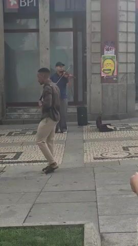 A street musician plays the violin on a city sidewalk. He is dressed in a blue shirt, grey pants, and a baseball cap. A black speaker and an open violin case are on the ground near him. A man in a brown shirt and tan pants stands facing the musician, listening. Another man in a light blue shirt and jeans walks past in the foreground. The scene is set against the backdrop of a building with large windows.