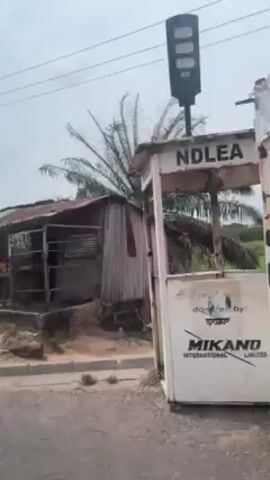 The video shows a view from a moving vehicle, likely a car, driving past a roadside structure and a motorcycle. The structure appears to be a small, simple shop or stall constructed of wood and corrugated metal, with a faded and partially deteriorated roof. Merchandise such as boxes and snacks are visible on a table in front of the stall. The ground around the structure is sandy. A palm tree is visible behind the building. A motorcycle carrying three people drives in the opposite direction along the road. The riders are wearing casual clothing and no helmets. The sky is overcast.