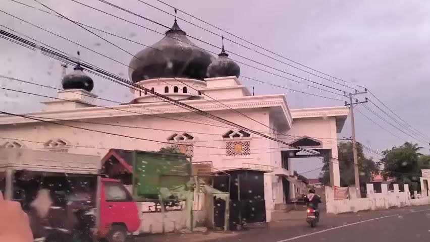 The video shows a white mosque with black domes, taken from inside a car on a rainy day. A person is holding a mobile phone, presumably to record the video, which shows the mosque from a moving vehicle. There are power lines crisscrossing the view. A motorbike drives by the mosque. The mosque has decorative windows and arches. The sky is overcast.