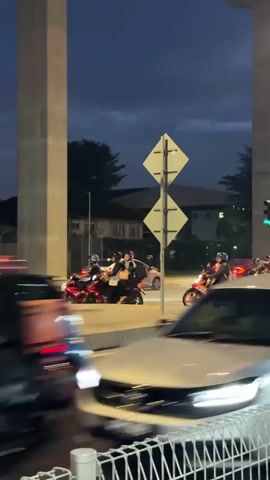 The video shows a street scene at dusk or early night, with a traffic light visible and illuminated. Several motorbikes are stopped on the side of the road, some with multiple riders. There are also cars in motion on the road. The scene is framed by large concrete pillars, suggesting an overpass or elevated structure. The sky is dark with some clouds, and the overall lighting is dim, indicating twilight or nighttime conditions.