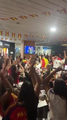 A crowded indoor space, possibly a bar or restaurant, is filled with people wearing red and yellow, the colors of the Spanish flag. Many are holding or waving Spanish flags and some are wearing Spanish flag-themed clothing or face paint. The crowd is cheering, raising their arms in celebration, and some are holding up their phones. A large screen displaying a soccer game is visible in the background, with a player in red and yellow on the screen. Small Spanish flags are strung across the ceiling. The atmosphere is one of excitement and collective jubilation, likely during a sports event.