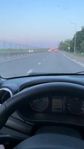 The video shows a view from inside a car driving on a multi-lane highway. The first shot is from the perspective of a front passenger, looking out the front windshield. The second shot is a close-up of the driver's side mirror, showing a sunset reflecting in it as other cars drive by.