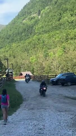 A woman wearing a pink top and grey shorts walks away from the camera on a gravel path. Two motorcycles are driving on the same path towards the viewer. The first motorcycle is closer to the viewer, and the second motorcycle is further away. Both motorcycles are ridden by people wearing dark clothing and helmets. In the background, there are parked cars, a wooden fence, and a forested mountain.