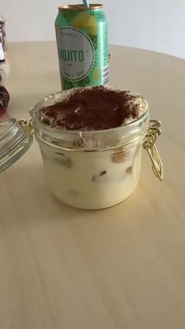 A close-up shot shows a tiramisu dessert in a glass jar with a clamp lid on a wooden table. The tiramisu is topped with cocoa powder. To the left of the jar, a green can with "Mojito" written on it is visible. The lid of the jar is partially open. The background is blurred.