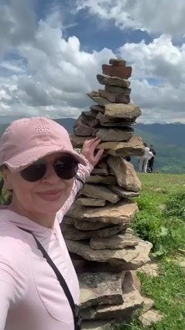 A woman in a pink baseball cap and sunglasses is taking a selfie in front of a tall stack of stones on a grassy mountainside. She is wearing a pink hoodie. Behind her, other people are visible on the mountain, some gathered around tables, and several vehicles are parked. The sky is cloudy with patches of blue. The environment appears to be a scenic, elevated location.