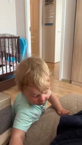 A blonde toddler wearing a light blue onesie with yellow and pink designs is on a brown textured surface, possibly a bed or couch. The toddler is facing towards the left side of the frame, looking downwards with a curious expression. Their right arm is extended forward, and their left arm is bent, resting on the surface. In the background, a wooden crib with bedding is visible on the left. A light blue object, possibly a toy, is propped against the crib. The room appears to be a bedroom, with a wooden floor and a door in the background that leads to another area of the house. A white cabinet is on the right side of the frame. A hand enters the frame from the top right and gently ruffles the toddler's hair.