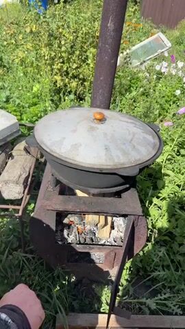 A medium shot shows a cast iron pot with a lid on top of a makeshift metal stove, outdoors. The stove is fueled by burning logs, with flames visible through an opening in the front. The pot is dark grey, and the lid is lighter grey with a wooden handle. The stove is rusted and appears to be made from repurposed metal. The background is a garden with green foliage and some flowering plants. The lighting suggests it is daytime, possibly late morning or early afternoon. A person's hand, wearing a plaid sleeve, is visible in the lower left corner, reaching towards the stove to adjust the logs. The camera is stationary.