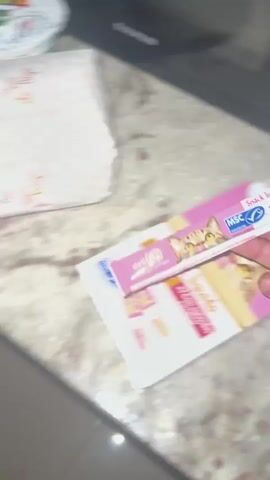 A person's hand holds a pink and white cat treat tube, featuring an illustration of a cat's face. In the background, a small tabby cat walks toward the hand holding the treat. The cat reaches the treat and begins to lick it. The environment appears to be indoors, with tiled flooring and a wooden door visible.