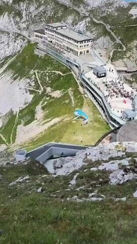 This aerial view shows a paraglider landing on a grassy slope next to a large building situated on a mountainside. Several people are visible on the building's terraces and walkways, observing the paraglider. Two paragliders, one blue and white, and another partially obscured, are on the ground, with at least three individuals near them. The environment is rocky and mountainous, with patches of green grass.