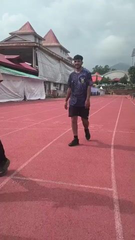 Two young men are walking on a red running track. The man on the left is wearing a tan t-shirt and black pants, and the man on the right is wearing a dark blue t-shirt with a circular pattern and dark shorts. They are walking in the same direction, away from the camera. In the background, there is a building with a red roof and white siding, and some trees. The sky is cloudy.