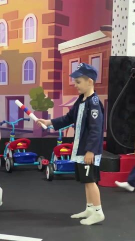 A young boy in a blue police uniform is playing traffic cop in what appears to be an indoor play area made to look like a city. The boy holds a baton with red and white stripes, and he is directing pedestrian traffic, waving the baton back and forth. In the background, a man in a white t-shirt stands near a road sign that shows stick figure children crossing the street. Behind the boy are several red and blue tricycles. Behind the tricycles, a mural depicts a cityscape. The boy is wearing white socks with black logos, and another child in a red jacket walks in the background. The floor is dark and marked to look like a street, complete with painted white lines. A yellow gasoline pump with "95" written on it can also be seen.