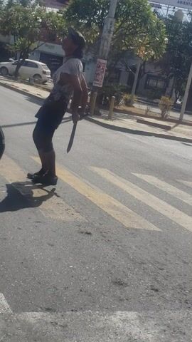 Here is a description of the video:
The video shows a man in a striped shirt, dark shorts, and a baseball cap doing push-ups on a crosswalk. He is using a long, thin object, possibly a stick or sword, to help him balance. Cars are parked nearby, and a billboard is visible in the background. The scene appears to be a street in a city.