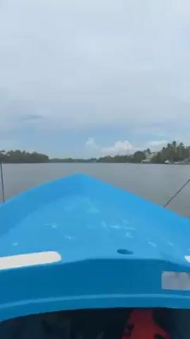The video shows a view from a blue motorboat moving across a body of water. The boat has a pointed bow and a visible rope extending from the front. On the horizon, the shoreline is lined with palm trees and buildings, some with thatched roofs. The sky is overcast with a mix of white and gray clouds. The water appears calm, reflecting the gray sky. An orange life jacket is visible inside the boat.