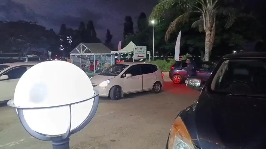 At night, the camera pans across a parking lot with several parked cars, revealing a woman walking toward the camera. The parking lot is lit by streetlights and round outdoor lamps.  A building with a sign that reads "Pharmacy" is visible in the background. The woman is wearing a white top and a black jacket. She looks up at the camera and then continues walking.  The camera movement is shaky.