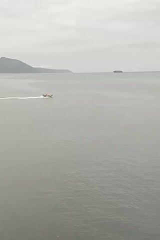 A motorboat is traveling across a body of water. The water is calm and appears to be gray. The sky is overcast and the clouds are also gray. In the distance, there are hills and a large ship. The motorboat is white and orange and has a wake behind it. The boat appears to be moving at a good speed. The overall scene is muted and somber.