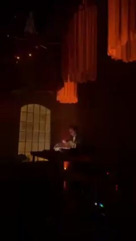 A man DJs in a dimly lit room with red lighting, with decorative fabric chandeliers and hanging star shapes overhead. He stands at a table with equipment, adjusting the controls. Behind him is a large arched window. There are other tables with equipment nearby. The video has a dark and grainy quality.