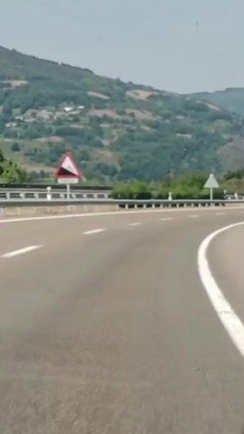 The video shows a view from a vehicle driving on a highway. The road curves to the right. On the left side of the road, there is a guardrail and a green, forested hillside with some buildings visible in the distance. On the right side of the road, there are more trees and a gentle slope. The sky is overcast.