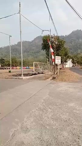The video pans across an outdoor area with a paved road in the foreground. In the background, there is a large open space with several small colorful vehicles, likely for rent or play, and people scattered around. A few trees are visible, and further in the distance, a gentle mountain slope covered in vegetation rises against a hazy sky. There are also some poles with wires strung between them, suggesting some form of overhead lighting or infrastructure. To the right, a white building and a striped pole resembling a barrier are visible.