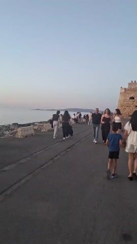 The video captures a street scene featuring a crowd of people walking towards a stone castle on the right side of the frame. The sky is clear, and the sea is visible on the left side of the street. The camera follows a group of people, including a child wearing a blue shirt and black shorts, who is holding hands with an adult. Various individuals and groups stroll along the street, dressed in casual clothing. The castle is made of light-colored stone and has a series of crenellations along its top edge. The road is paved and appears to be a popular walking area.