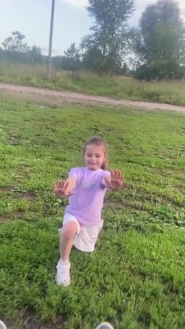 A young girl with blonde hair, wearing a purple t-shirt and white shorts, is performing lunges on a grassy field. Her arms are extended forward, and she has a determined smile on her face. Her white sneakers are visible as she moves. The perspective is from someone lying on the grass, looking up at the girl, with their own feet and shoes in the foreground. The background consists of a dirt path, trees, and a hint of a cloudy sky, suggesting it's daytime. The overall atmosphere is playful and active. No visible text is present.