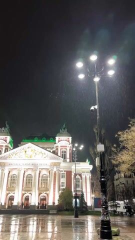 The video shows the Ivan Vazov National Theatre in Sofia, Bulgaria at night during a rainstorm. The building is brightly lit, with its red and white facade and green roof standing out against the dark sky. Rain is visible, creating a wet sheen on the square in front of the theater. A tall, ornate lamppost with multiple lights and a security camera is prominent in the foreground. Several people are seen walking near the theater entrance.