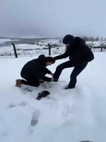 In a snowy outdoor setting, two individuals are interacting. One person, wearing a black puffy jacket and brown boots, is kneeling in the snow, appearing to tie or adjust something on the other person's boot. The second person, wearing a black puffy jacket, dark jeans, and a navy beanie, is standing, with one leg extended forward and the other planted in the snow. The standing person has their hand resting on the kneeling person's head, possibly for balance or support. There is a pair of gloves lying in the snow nearby. In the background, there is a wire fence, a body of water, and snow-covered landscape under a cloudy sky. Footprints are visible in the snow.