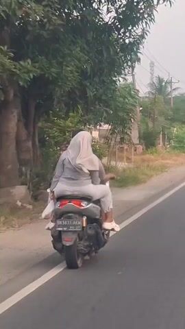 Two individuals are riding a motorcycle on a paved road. The person in the rear, who appears to be a woman wearing a light gray hijab and matching pants and top, is seated behind the driver. The driver's upper body is obscured. The motorcycle is traveling away from the camera. The environment appears to be rural or suburban, with trees and vegetation visible along the roadside. There are some buildings in the background. The weather seems to be overcast. The license plate of the motorcycle is partially visible.