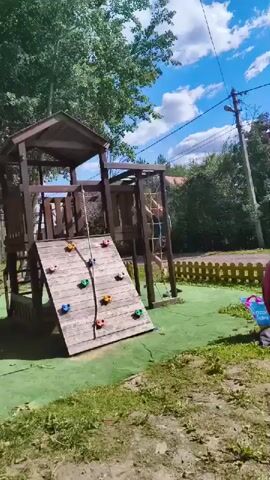 A child is climbing a rope ladder on a wooden playground structure. The playground has a climbing wall with colorful grips and a slide. The ground is covered in green artificial turf with some patches of grass and dirt around the edges. A person in a pink shirt is partially visible on the right side of the frame. The sky is blue with white clouds.