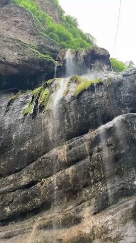A low-angle, static shot shows a waterfall cascading down a steep, rocky cliff face. The rock is dark and appears wet, with patches of green moss growing on its surface. The water flows in multiple streams, some wide and some thin, creating streaks of white against the dark rock. The top of the waterfall is obscured by the cliff and lush green vegetation, which spills over the edge. The sky above is overcast and bright. A thin wire or cable is visible extending diagonally across the upper right portion of the frame. The overall impression is one of a rugged, natural landscape.