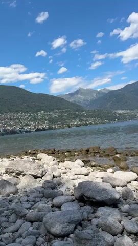 This is a vertical, outdoor, handheld video taken from a low angle, showing a rocky shoreline in the foreground and a body of water with mountains in the background. The sky is blue with scattered white clouds. The water appears calm, with small ripples. The shoreline is covered with numerous smooth, gray and brown rocks of various sizes. In the distance, there is a town or city nestled along the edge of the water, with buildings visible on a green, sloping mountainside. The mountains themselves are tall and covered in vegetation, with some rocky peaks visible at the highest elevations. The overall impression is a serene, natural landscape under a bright, sunny sky. There are no people or animals visible in the video. The camera is mostly stationary, with only minor movements that appear to be the result of handholding.