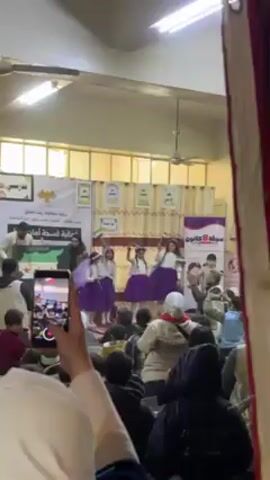 The video shows a group of young girls dancing on a stage during an event. The girls are wearing white tops and purple skirts, with floral headbands. Behind them are banners with Arabic writing and images, likely indicating the event or organization hosting it. A few adults are on stage with the girls, possibly supervising or coordinating the performance. The audience consists of children and adults, some of whom are recording the performance with their phones. The setting appears to be a school gymnasium or auditorium with fluorescent lighting and beige walls.