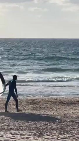 A person in a dark wetsuit stands on a sandy beach, holding a kite or sail. The kite is mostly white with black geometric designs and the number "10" visible. The person faces the ocean, which has moderate waves under a partly cloudy sky. They take a few steps while holding the kite.