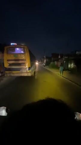The video displays a first-person point-of-view from a vehicle, likely a motorcycle, traveling at night. The vehicle's handlebars are visible at the bottom of the frame, and a digital speed display briefly shows "35." A bright yellow beam of light illuminates the wet road ahead. On the left side of the road, a large yellow bus with illuminated purple interior lights is stationary or moving slowly in the same direction as the camera. On the right side of the road, a person walks away from the camera. Later, another individual riding a light-colored scooter or motorcycle passes on the right, traveling in the opposite direction. The sky is dark, and streetlights are visible overhead along the road, which appears to be a two-lane thoroughfare with a broken white line. Buildings line the right side of the road in the distance.
