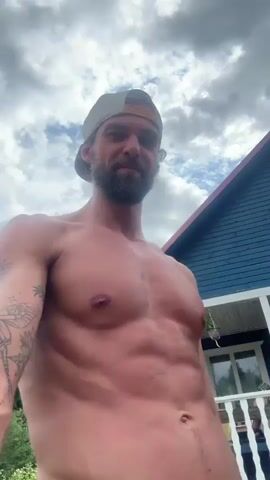 The video shows a shirtless man with a beard, wearing a baseball cap, outdoors. He has tattoos on his left arm, including a butterfly design. The man is looking around and appears to be in a rural setting, with a wooden building and trees visible in the background. The sky is partly cloudy, suggesting it is daytime. The man's physique is prominent, and he seems to be in good physical condition. There is no audible sound in the video, and no visible text. The setting appears to be Kuti, Russia, as indicated by the provided location context. The overall mood is relaxed and casual, with the man enjoying the outdoors.