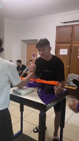 Two people are arm wrestling at a table with an arm wrestling setup. They are inside a room with tiled floors. Several other people are sitting on chairs, watching. There are various flags hanging from the ceiling. A sign on a closed door reads "CUISINE".