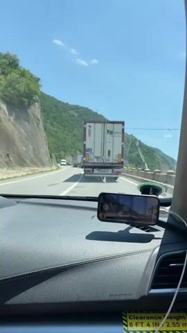 The video captures a scenic drive along a lake with mountains in the background. The first frame shows the driver's perspective from inside a car, with a truck ahead and a smartphone mounted on the dashboard. The following frames, taken from the passenger's side, showcase the lake's expanse, flanked by rolling green mountains under a bright blue sky with fluffy white clouds. The car's side mirror and a concrete barrier are visible in the foreground, framing the tranquil landscape. The video highlights the picturesque journey, emphasizing the natural beauty of the lake and mountains.