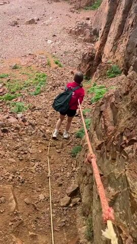 A young woman in a red sweatshirt and black shorts descends a rocky, steep slope towards a beach. She is holding onto a thick rope, likely for support, as she navigates the uneven terrain. The slope is composed of reddish-brown rocks and sparse vegetation. To the right, a sheer cliff face rises, also made of red rock. The beach below is a similar reddish-brown color and extends towards the ocean. In the distance, a rocky islet can be seen in the water. The sky is overcast, suggesting a cloudy day.