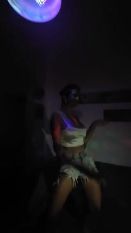 A woman is dancing in a dimly lit room with a disco light. She is wearing a white top and light-blue jean shorts, and she has her hair pulled back. She dances with a wide smile and energetic movements. There is a couch in the background, and some wall decorations are visible. The room appears to be a living space.