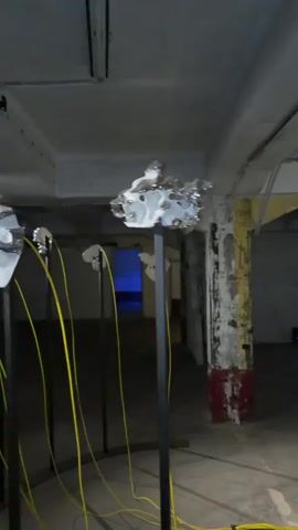 The video shows an art installation in a dimly lit, industrial-looking space. Several white, mask-like sculptures with metallic, textured elements are displayed on tall black stands. Yellow cables hang from these sculptures and trail onto the floor. In the background, a screen shows a person in blue, and a doorway with blue light can be seen. The walls are concrete and show signs of wear.